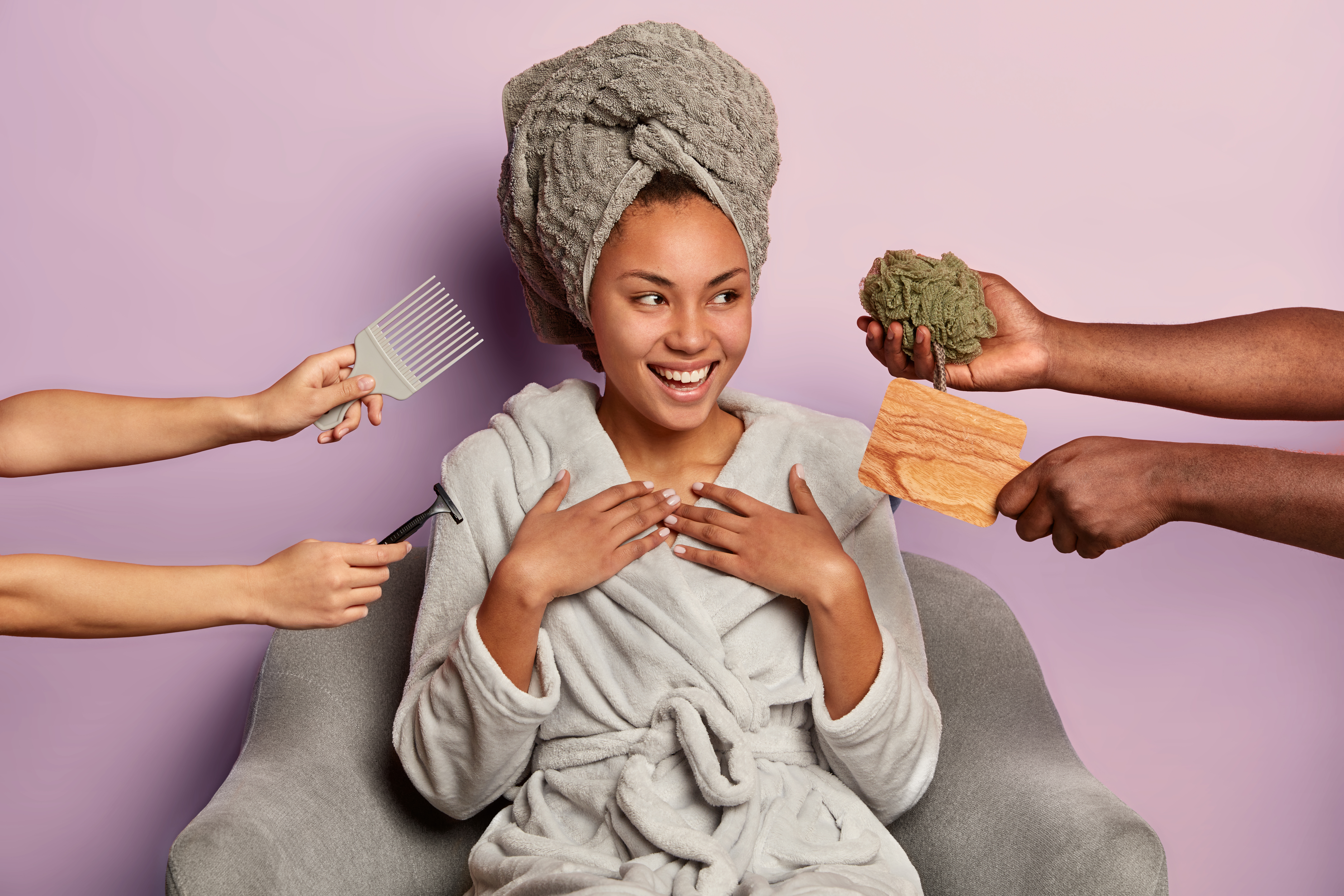 Photo of satisfied relaxed dark skinnned woman wears wrapped towel and bathrobe after shower, surrounded by hands with razor, mirror, sponge and comb, has pleasant smile, isolated on purple background